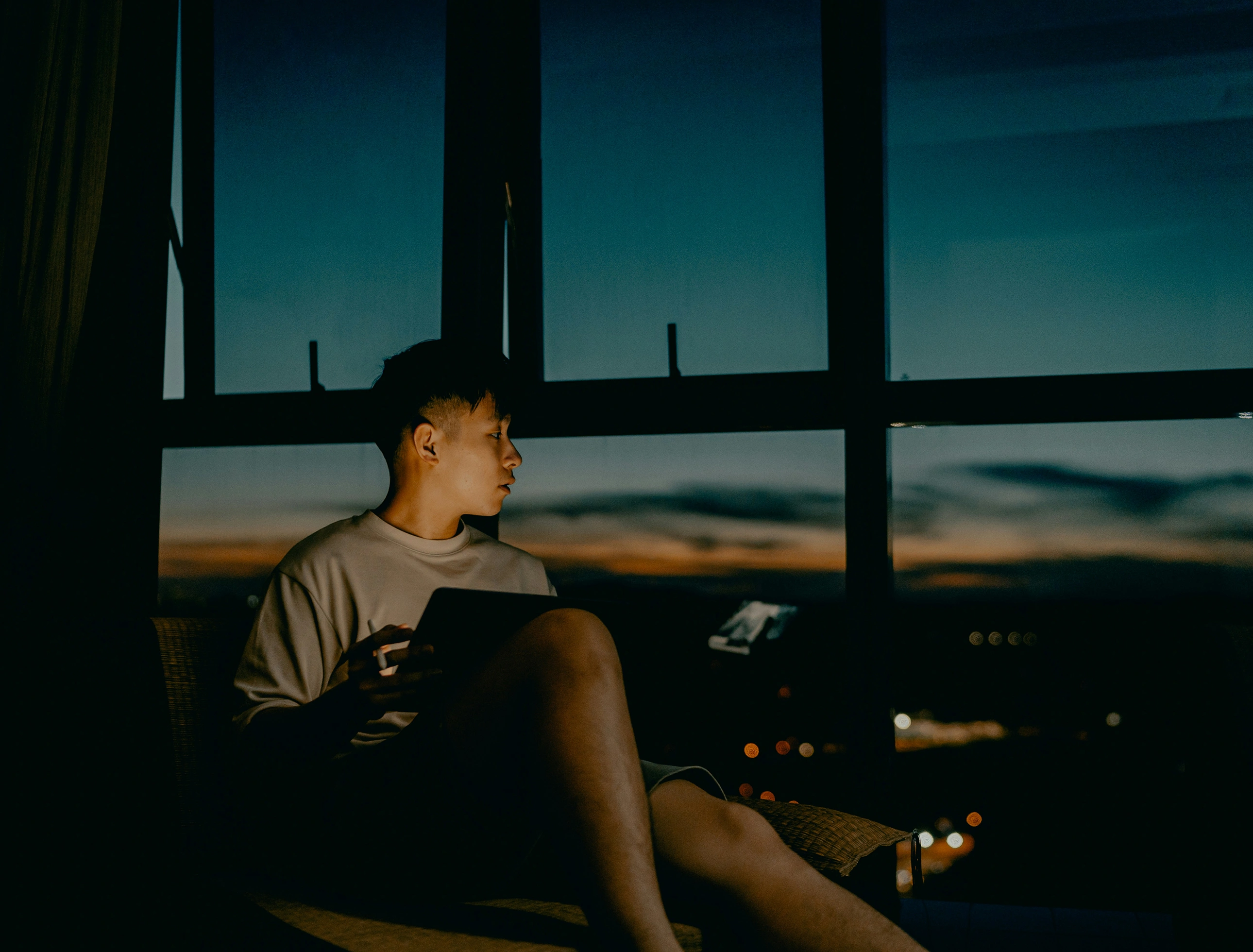 A man sitting and looking out a large window in the late evening. There is a tablet in his lap which illuminates his face, and blurred streetlights can be seen outside. 