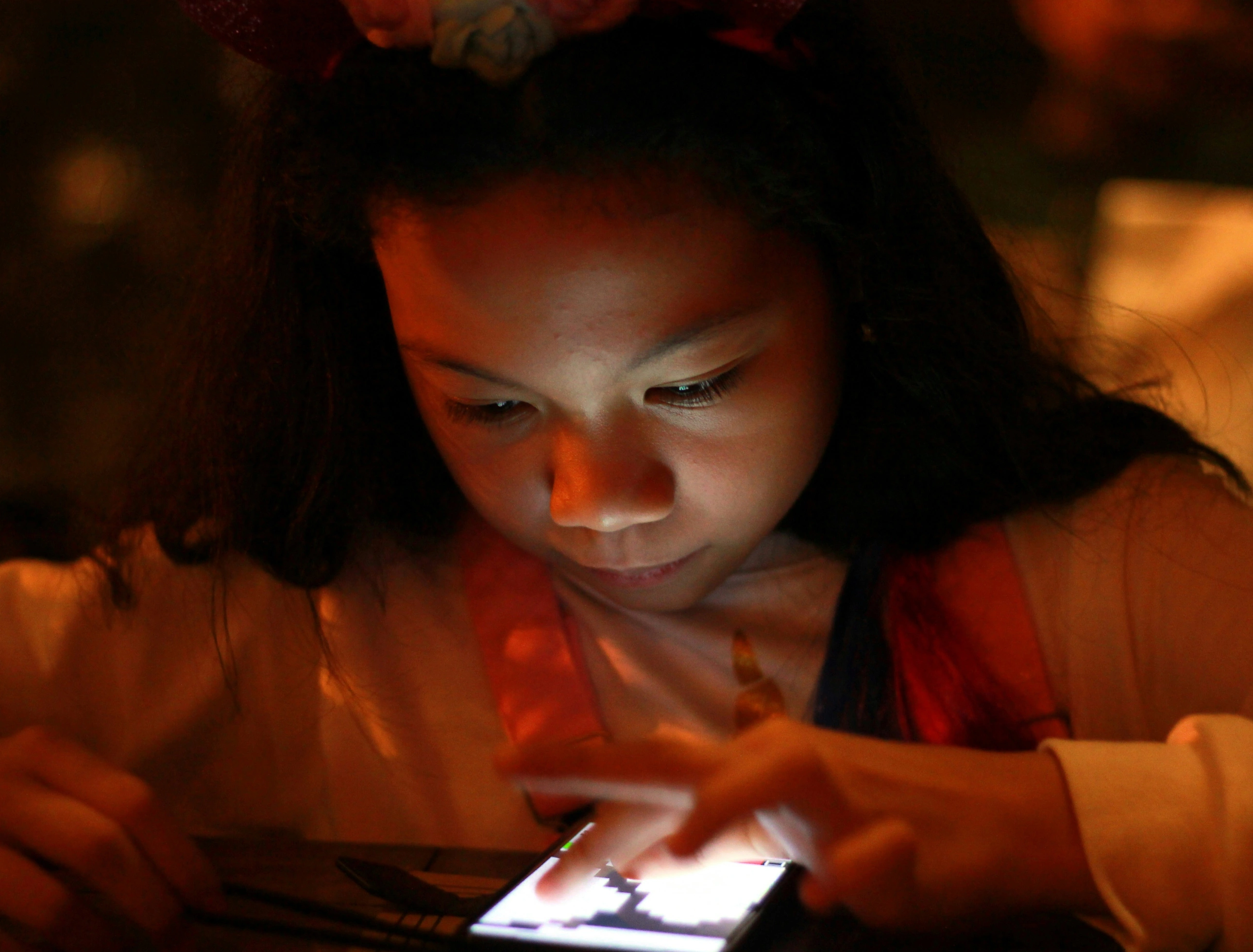 A young girl scrolling on a phone, the light illuminating her face. The background is soft, orange and out of focus.