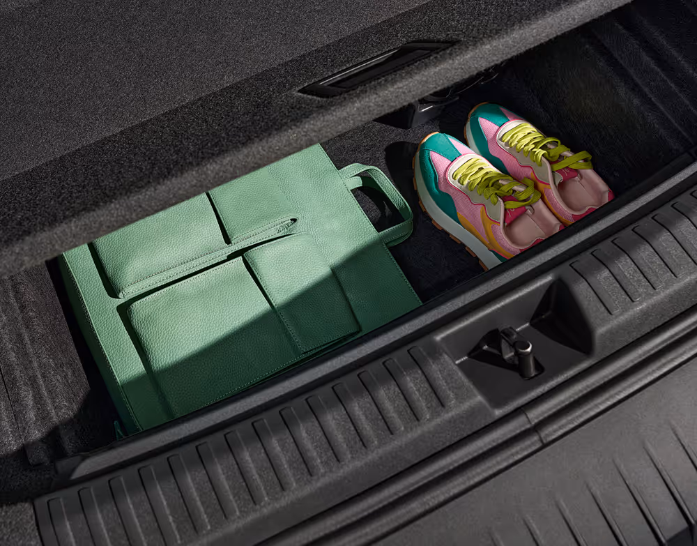 Blick auf den leicht angehobenen Ladeboden im Kofferraum des Škoda Epiq, der ein zusätzliches Unterfach freigibt. In diesem versteckten Stauraum direkt hinter der Ladekante sind eine hellgrüne Tasche und ein Paar bunte Sneaker verstaut. Die Detailaufnahme veranschaulicht die praktische Nutzung des zusätzlichen Platzangebots.