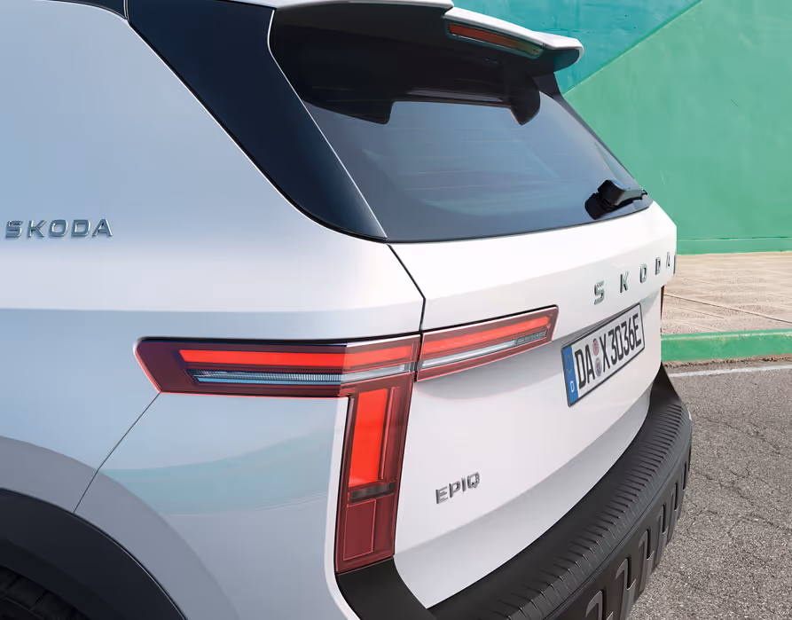 Eine Nahaufnahme zeigt die Heckpartie und die Fahrzeugflanke des Škoda Epiq in der Lackierung Platinum-Weiß Metallic. Im Fokus stehen die markanten, T-förmigen LED-Rückleuchten, der schwarze Dachspoiler sowie die Škoda Schriftzüge auf der Heckklappe und der C-Säule. Ein robuster, dunkler Stoßfänger schließt das Fahrzeug nach unten ab, während im Hintergrund eine grüne Gebäudewand zu sehen ist.