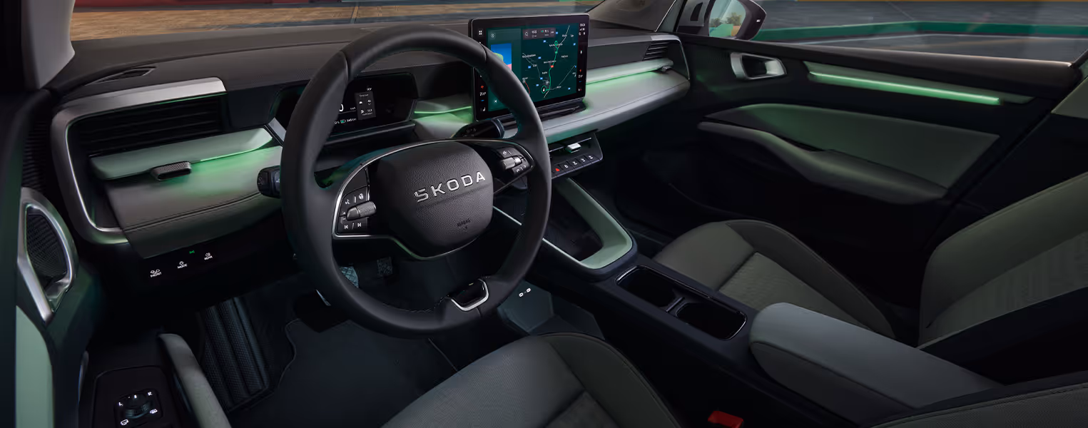 Eine leicht erhöhte, schräge Innenansicht zeigt das Cockpit des Škoda Epiq. Im Fokus stehen das Zweispeichen-Multifunktionslenkrad, ein kompaktes digitales Kombiinstrument sowie der freistehende Infotainmentbildschirm auf dem aufgeräumten Armaturenbrett. Eine dezente grüne Ambientebeleuchtung, helle Zierelemente an den Türverkleidungen und graue Stoffsitze runden das schlichte Interieur ab.