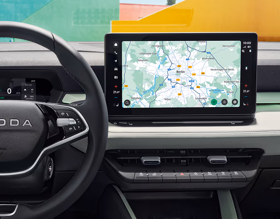 Eine Nahaufnahme des Cockpits im Škoda Epiq rückt den großen, freistehenden Infotainmentbildschirm mit einer aktiven Navigationskarte in den Fokus. Unterhalb des Displays befinden sich physische Bedientasten sowie Lüftungsauslässe, während am linken Bildrand das Multifunktionslenkrad und das digitale Kombiinstrument angeschnitten sind. Durch die Windschutzscheibe ist im Hintergrund eine farbenfrohe Architektur mit grünen und gelben Flächen erkennbar.
