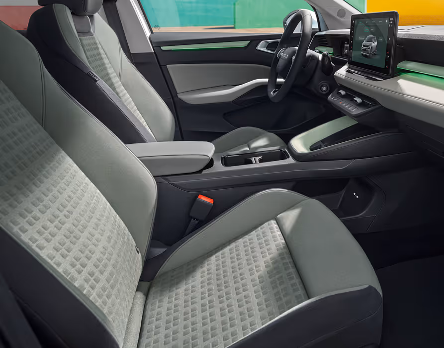 Eine seitliche Innenansicht des Škoda Epiq zeigt das Interieur der Design Selection Loft Mint. Die mintfarbenen Textilsitze mit korrespondierenden Nähten sowie die Armaturentafel mit grauem Techtona und matten Dekorelementen sind zu sehen, während durch die Fenster farbenfrohe, geometrische Gebäudewände sichtbar sind.