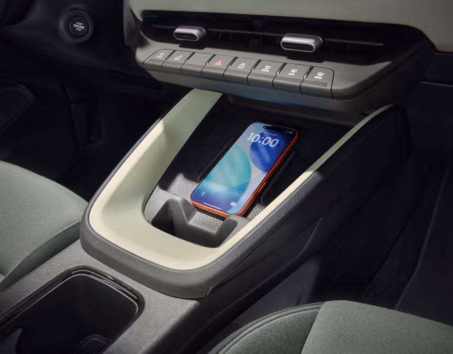 Eine Nahaufnahme der Mittelkonsole im Škoda Epiq zeigt ein vertieftes Ablagefach für Smartphones. In der mit einer hellen Zierleiste umrandeten Ladeschale liegt ein Mobiltelefon mit beleuchtetem Bildschirm. Oberhalb des Fachs sind eine Reihe physischer Bedientasten und Lüftungsauslässe sowie links am Armaturenbrett der Start-Stopp-Knopf zu erkennen.
