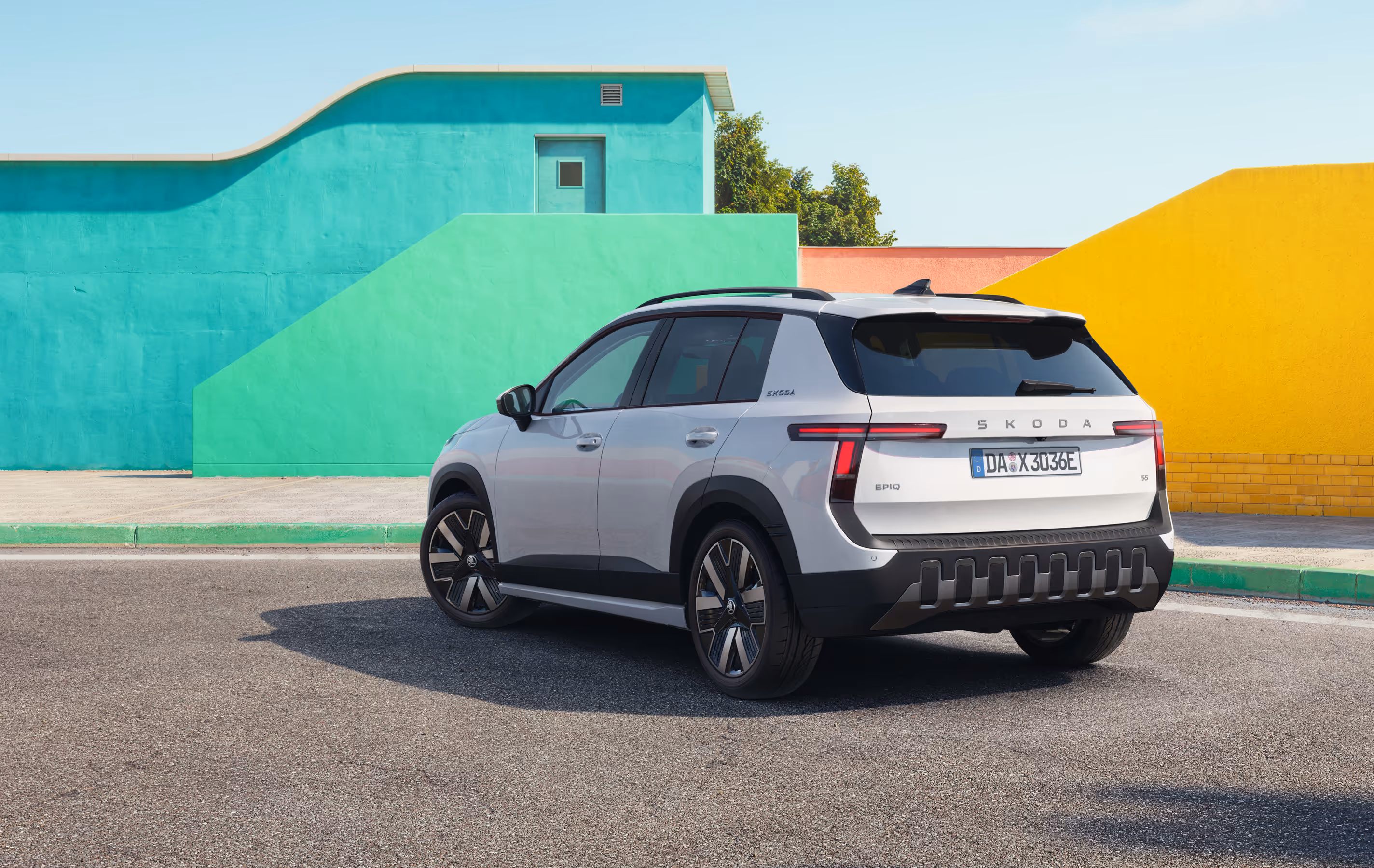 Ein geparkter Škoda Epiq in der Lackierung Platinum-Weiß Metallic steht auf einer asphaltierten Fläche. Die Ansicht von schräg hinten zeigt markante, T-förmige LED-Rückleuchten, auffällige Leichtmetallfelgen sowie einen robusten, dunklen Stoßfänger mit vertikalen Elementen. Im sonnigen Hintergrund bilden geometrische Gebäudewände einen farbenfrohen Kontrast zum Fahrzeug.