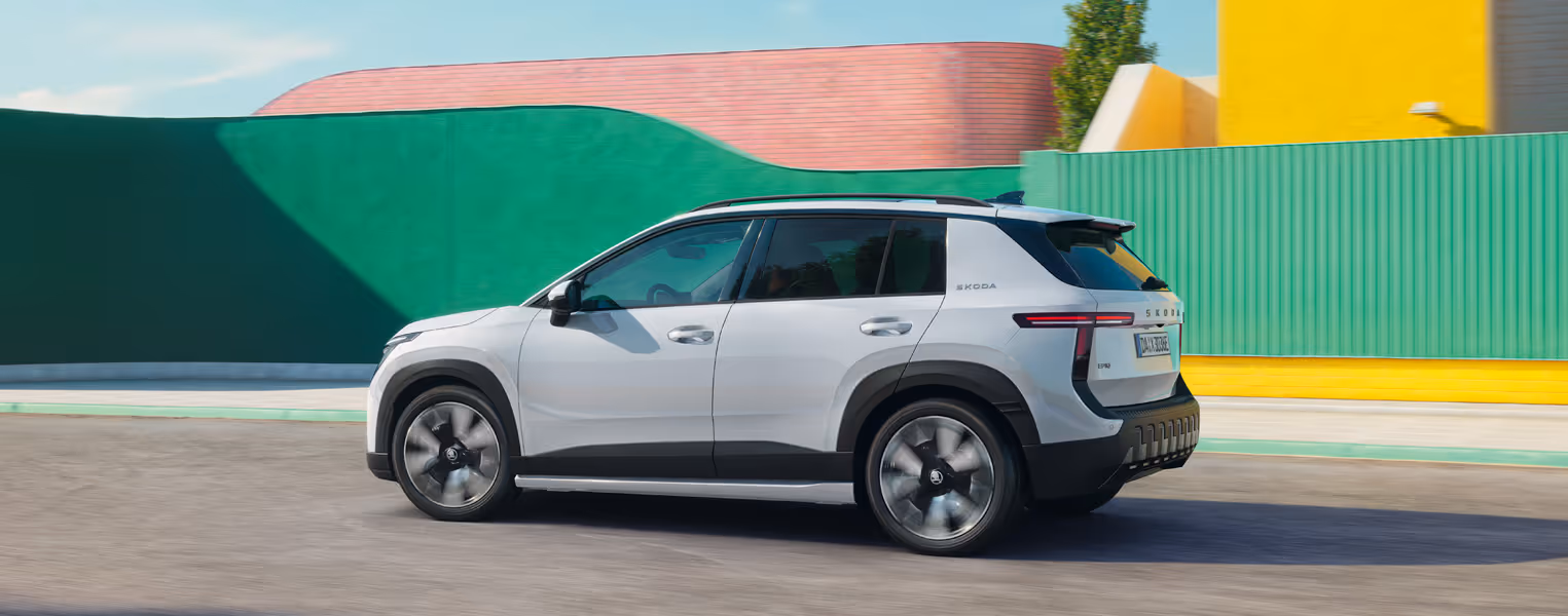 Ein Škoda Epiq in der Lackierung Platinum-Weiß Metallic fährt auf einer asphaltierten Straße. Die dynamische Seitenansicht zeigt dunkle Kontrastelemente an den Radkästen und Seitenschwellern, markante T-förmige Rückleuchten sowie rotierende Leichtmetallfelgen, die die Bewegung unterstreichen. Im sonnigen Hintergrund zieht eine moderne Architektur mit farbenfrohen, geometrischen Gebäudewänden vorbei.