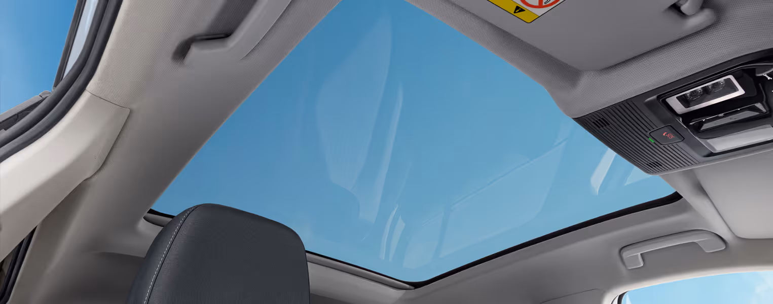 Blick nach oben im Innenraum des Škoda Epiq auf ein großflächiges Panorama-Glasdach, durch das ein blauer Himmel zu sehen ist. Am hellgrauen Dachhimmel sind seitliche Haltegriffe, eine Sonnenblende sowie die vordere Bedieneinheit mit integrierter SOS-Taste angebracht. Im unteren Bildrand ist der obere Teil einer dunklen Sitz-Kopfstütze mit hellen Ziernähten erkennbar.