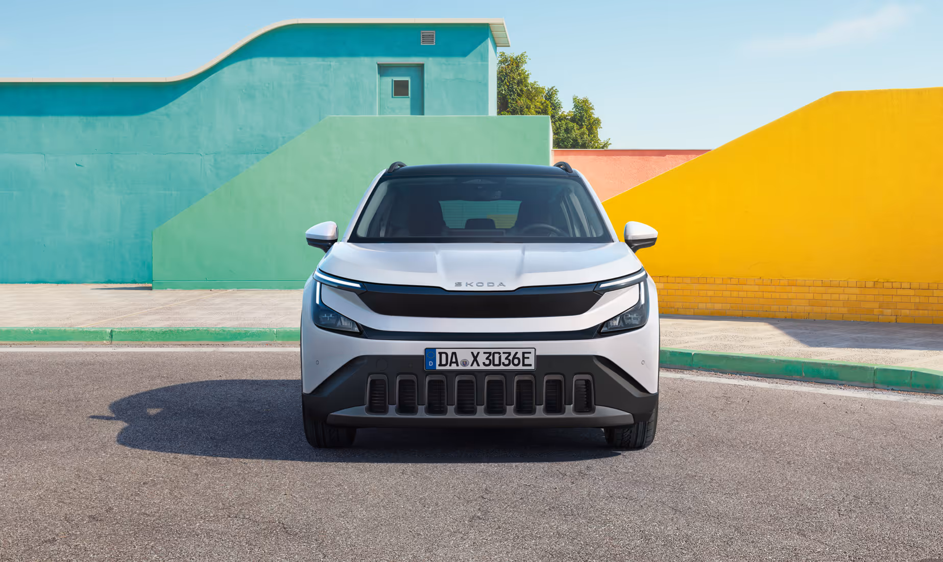 Frontansicht des Škoda Epiq in Platinum-Weiß Metallic, der auf einer asphaltierten Fläche parkt. Im Hintergrund heben sich geometrische Wände in Pastellgrün und Gelb kontrastreich gegen einen strahlend blauen Himmel ab. Die Fahrzeugfront wird durch schmale LED-Scheinwerfer, eine durchgehende schwarze Blende und einen markanten Stoßfänger mit vertikalen Einlässen geprägt.