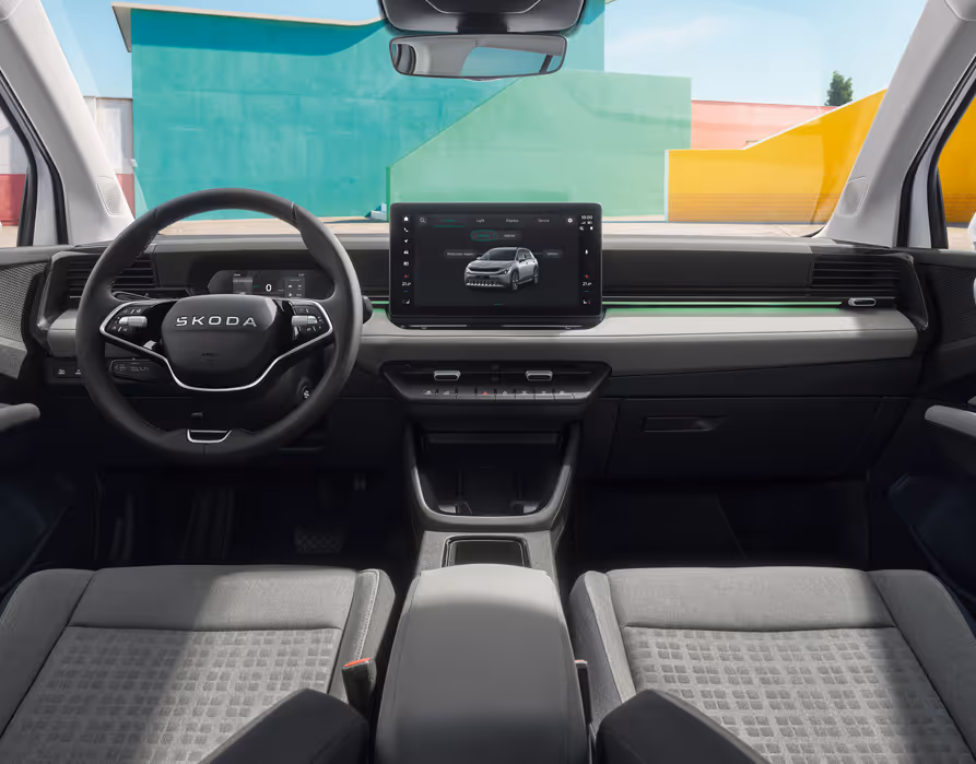 Eine weite Innenansicht zeigt das gesamte Cockpit des Škoda Epiq mit der Design Selection Loft Grau. Das Interieur zeichnet sich durch graue Textilsitze mit dezenten Ziernähten, mattes Dekor und graues Techtona an der Armaturentafel aus, während das Multifunktionslenkrad und der Infotainmentbildschirm zentral angeordnet sind. Durch die Windschutzscheibe sind farbenfrohe, geometrische Gebäudewände zu sehen.