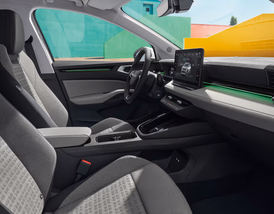 Eine seitliche Innenansicht des Škoda Epiq zeigt das Interieur der Design Selection Loft Grau. Die grauen Textilsitze mit dezenten Ziernähten sowie die Armaturentafel mit grauem Techtona und matten Dekorelementen sind zu sehen, während durch die Fenster farbenfrohe, geometrische Gebäudewände sichtbar sind.