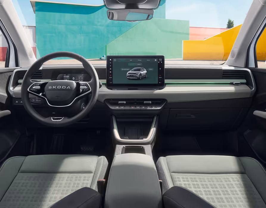 Eine weite Innenansicht zeigt das gesamte Cockpit des Škoda Epiq mit der Design Selection Loft Mint. Das Interieur zeichnet sich durch mintfarbene Textilsitze mit korrespondierenden Nähten, mattes Dekor und graues Techtona an der Armaturentafel aus, während das Multifunktionslenkrad und der Infotainmentbildschirm zentral angeordnet sind. Durch die Windschutzscheibe sind farbenfrohe, geometrische Gebäudewände zu sehen.