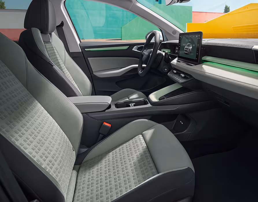 Eine seitliche Innenansicht des Škoda Epiq zeigt das Interieur der Design Selection Loft Mint. Die mintfarbenen Textilsitze mit korrespondierenden Nähten sowie die Armaturentafel mit grauem Techtona und matten Dekorelementen sind zu sehen, während durch die Fenster farbenfrohe, geometrische Gebäudewände sichtbar sind.