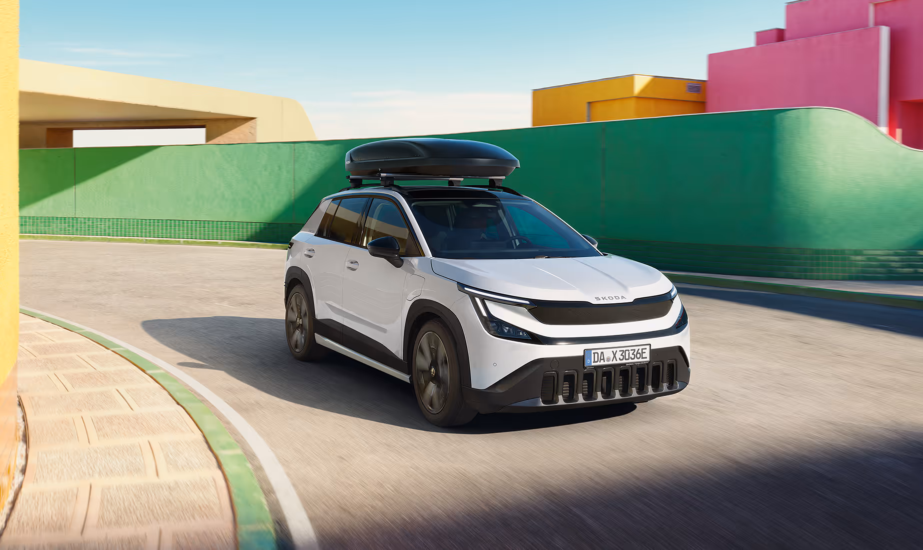 Dynamische Fahrszene des Škoda Epiq in der Farbe Platinum-Weiß Metallic, der mit einer großen, schwarzen Dachbox ausgestattet ist. Im Hintergrund hebt sich das Fahrzeug kontrastreich von bunten, geometrischen Mauern ab.