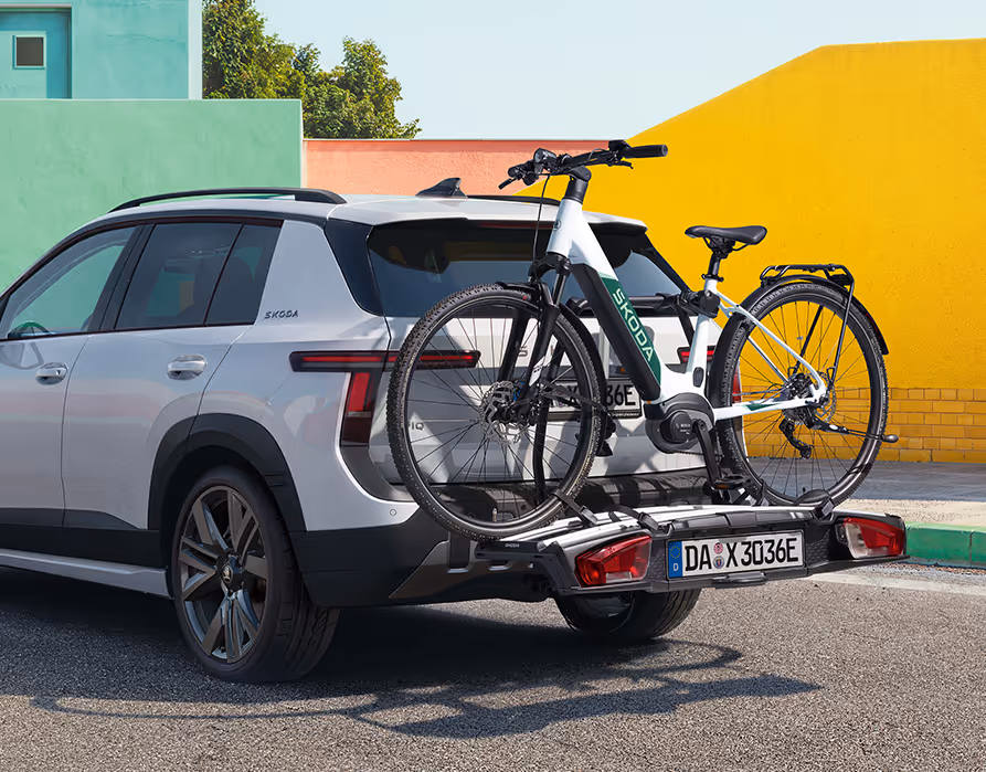 Heck- und Seitenansicht des Škoda Epiq in Platinum-Weiß Metallic, der mit einem beladenen Fahrradträger auf einer asphaltierten Fläche steht. Auf dem Träger ist ein helles E-Bike befestigt, das vor der modernen Heckpartie mit robustem, dunklem Stoßfänger platziert ist. Den Hintergrund bilden geometrische Wände in Pastellgrün und leuchtendem Gelb unter einem blauen Himmel.