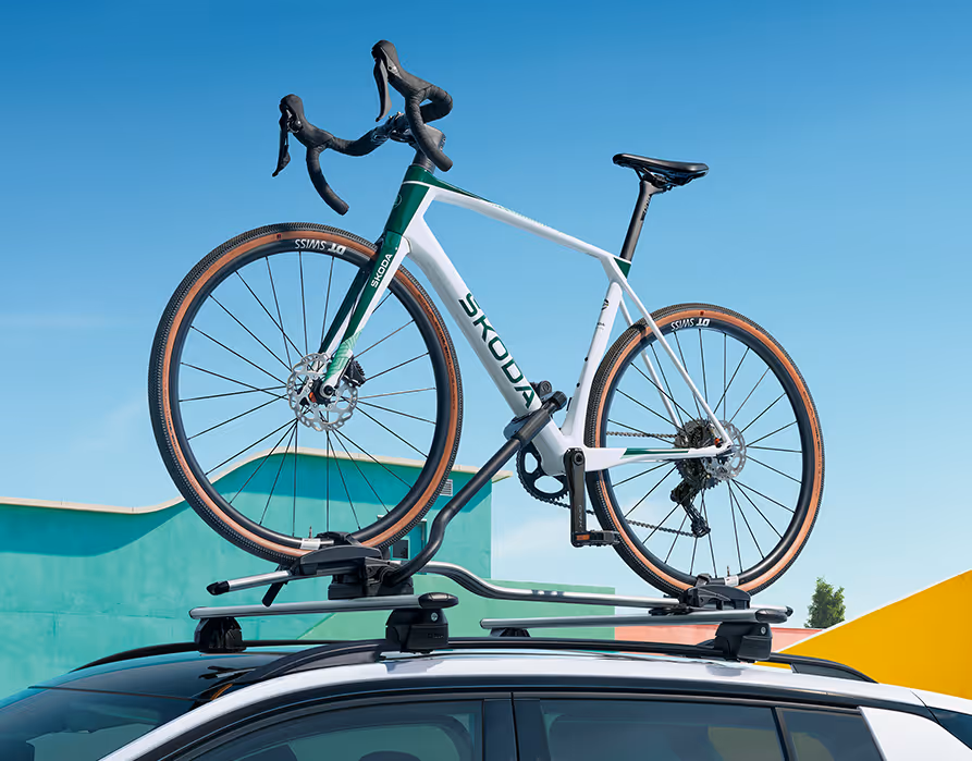 Detailaufnahme des Daches eines Škoda Epiq in Platinum-Weiß Metallic, das mit einem robusten Dachträger-System ausgestattet ist. Auf dem Träger ist ein weiß-grünes Rennrad sicher für den Transport fixiert. Diese Ansicht demonstriert die praktische Möglichkeit, sperrige Sportausrüstung platzsparend auf dem Fahrzeugdach zu transportieren.
