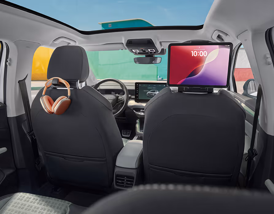 Blick aus dem Fond des Škoda Epiq auf die dunklen Rücklehnen der Vordersitze, die an den Kopfstützen mit sogenannten Smart-Holder-Aufsätzen ausgestattet sind. An der linken Halterung ist ein Kopfhörer aufgehängt, während auf der rechten Seite ein Tablet als Bildschirm für die hinteren Passagiere eingespannt ist. Im oberen Bildbereich ist das großzügige Panorama-Glasdach zu erkennen, durch das viel Tageslicht in den Innenraum fällt.