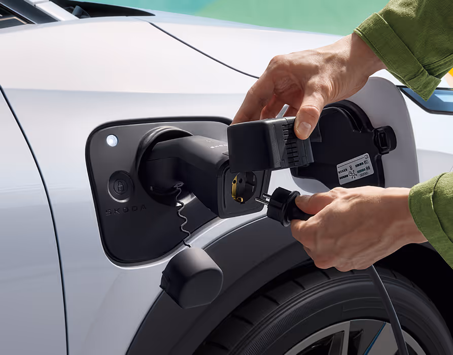 Detailaufnahme des geöffneten Ladeanschlusses am vorderen Kotflügel des Škoda Epiq in Platinum-Weiß Metallic. Die Hände einer Person stecken einen handelsüblichen Stromstecker in einen speziellen V2L-Adapter, der mit der Ladebuchse des Fahrzeugs verbunden ist.