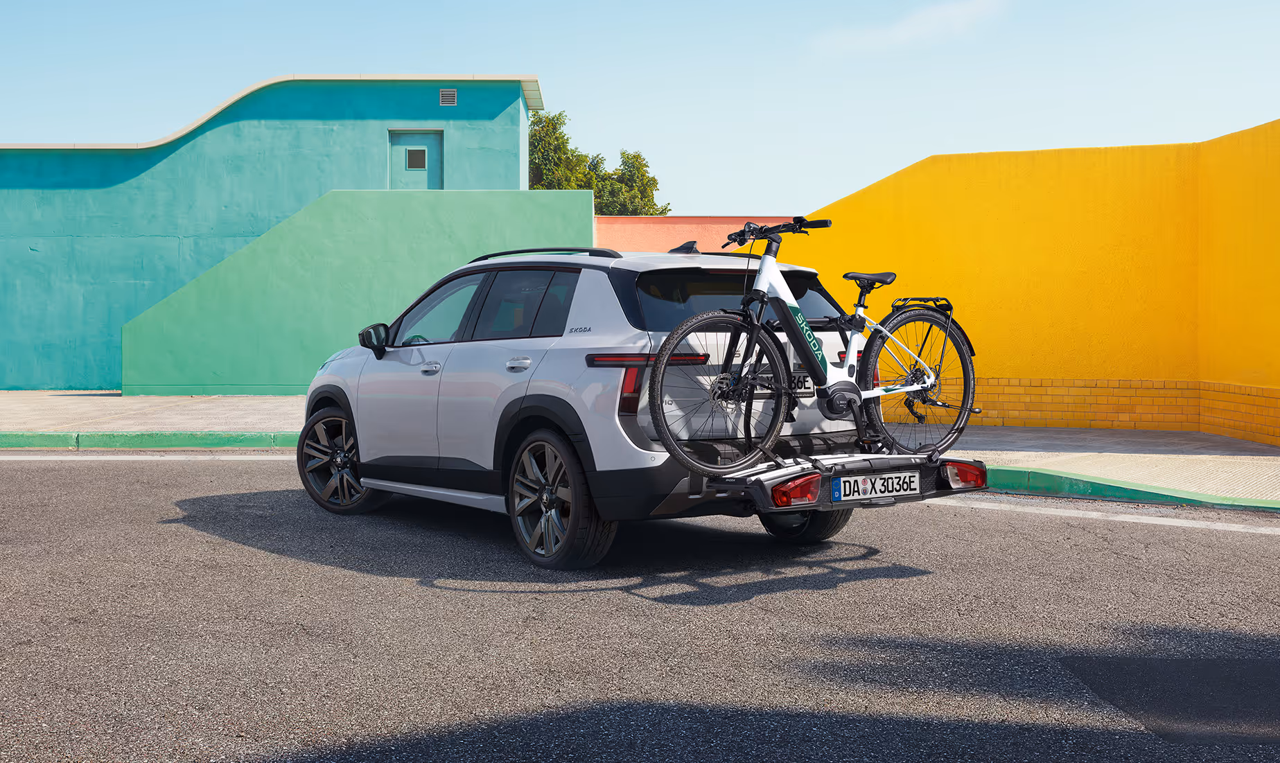 Heck- und Seitenansicht des Škoda Epiq in Platinum-Weiß Metallic, der mit einem beladenen Fahrradträger auf einer asphaltierten Fläche steht. Auf dem Träger ist ein helles E-Bike befestigt, das vor der modernen Heckpartie mit robustem, dunklem Stoßfänger platziert ist. Den Hintergrund bilden geometrische Wände in Pastellgrün und leuchtendem Gelb unter einem blauen Himmel.