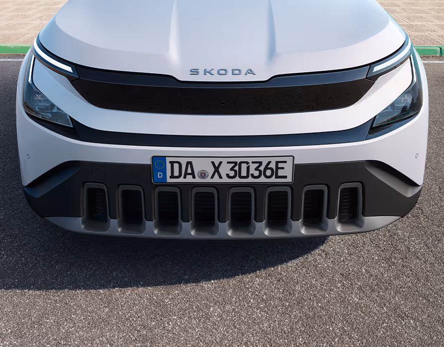 Eine frontale Nahaufnahme zeigt die markante Frontpartie des Škoda Epiq in der Lackierung Platinum-Weiß Metallic. Unterhalb des Škoda-Schriftzugs auf der Motorhaube befindet sich eine geschlossene, schwarze Blende, die die beiden LED-Scheinwerfer miteinander verbindet. Im unteren Bereich dominiert ein robuster, dunkler Stoßfänger mit einem mittigen Kennzeichen und auffälligen vertikalen Einlässen, der von einem silberfarbenen Element abgeschlossen wird.
