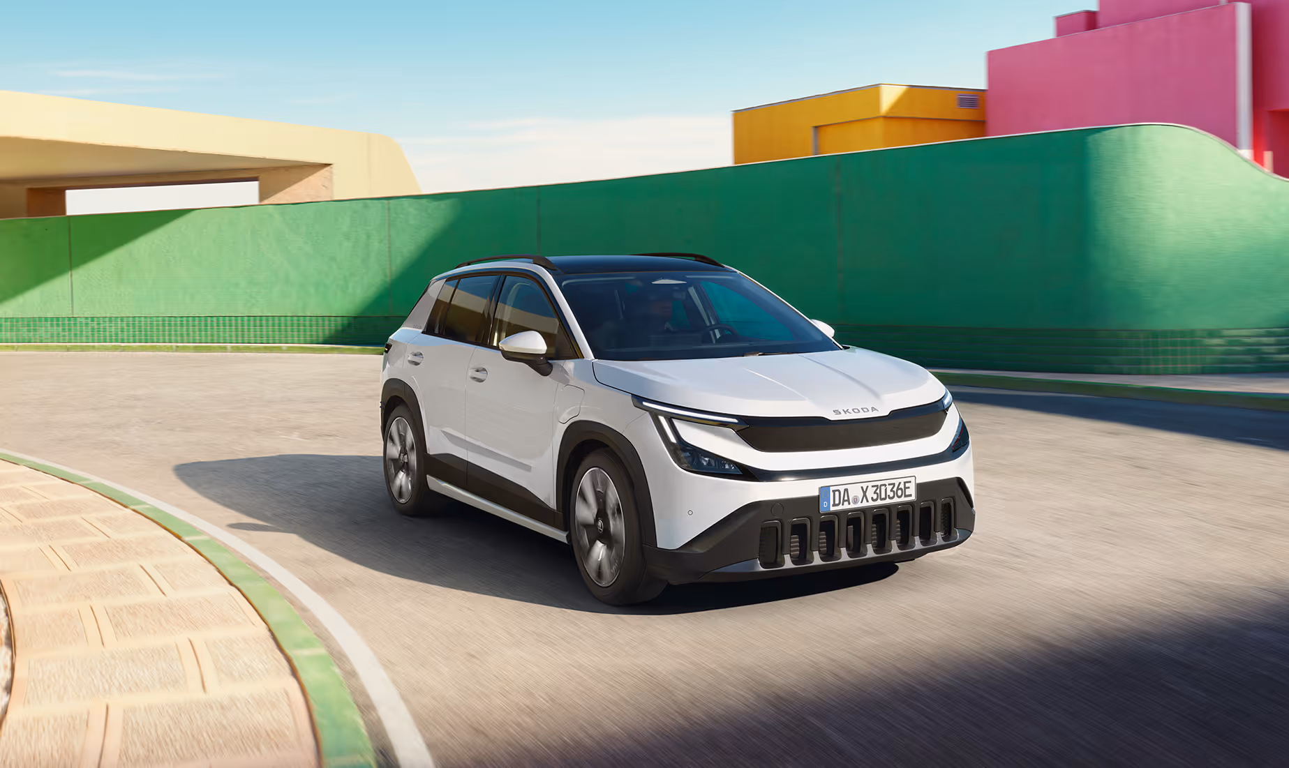 Ein Škoda Epiq in der Lackierung Platinum-Weiß Metallic fährt auf einer asphaltierten Straße. Das kompakte SUV-Crossover zeigt markante Designmerkmale wie dunkle Stoßfänger und eine geschlossene, schwarze Frontpartie. Im sonnigen Hintergrund ist eine moderne Architektur mit farbenfrohen, abstrakten Gebäudewänden in Grün, Gelb und Pink zu sehen.