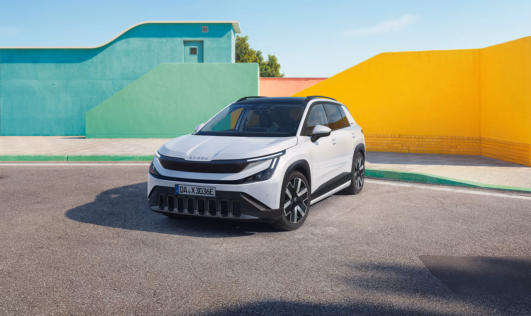 Ein geparkter Škoda Epiq in der Lackierung Platinum-Weiß Metallic steht auf einer asphaltierten Fläche. Die Ansicht von schräg vorne zeigt die geschlossene, schwarze Frontpartie, markante LED-Scheinwerfer sowie dunkle Leichtmetallfelgen und schwarze Seitenschweller. Im sonnigen Hintergrund bilden geometrische, farbige Wände in Grün, Rosa und leuchtendem Gelb einen starken Kontrast zum Fahrzeug.