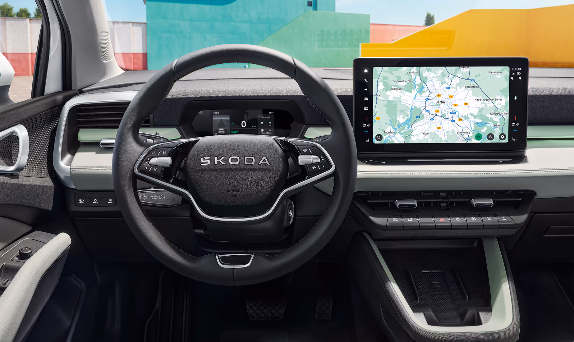 Eine Frontalansicht zeigt das moderne Cockpit des Škoda Epiq mit einem Zweispeichen-Multifunktionslenkrad, das den Škoda-Schriftzug trägt, und einem dahinterliegenden, kompakten digitalen Kombiinstrument. Zentral auf dem hell gestalteten Armaturenbrett befindet sich ein großer, freistehender Infotainmentbildschirm mit aktiver Navigationskarte, unter dem physische Bedientasten und eine Mittelkonsole mit Ablagefach angeordnet sind. Durch die Windschutzscheibe sind im Hintergrund farbenfrohe, geometrische Gebäudewände sichtbar.