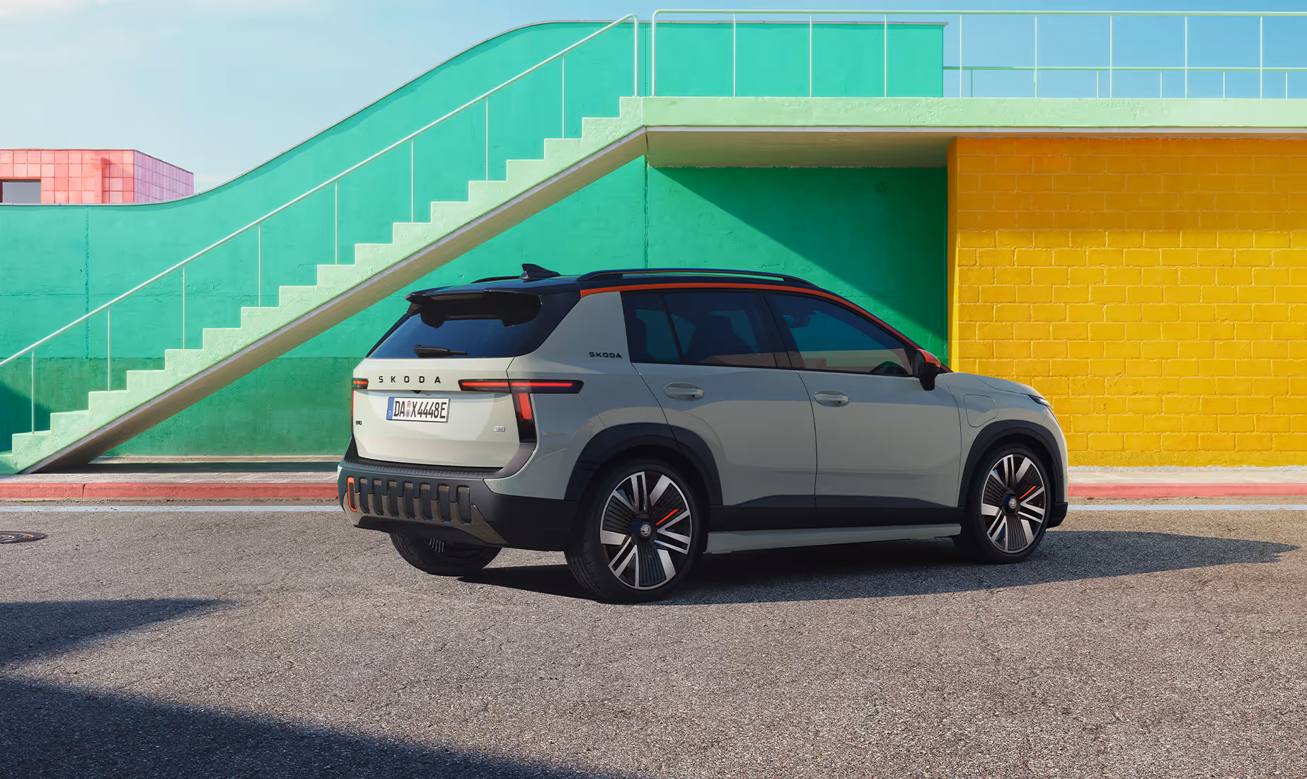 Heck- und Seitenansicht des Škoda Epiq als Sondermodell "First Edition", das auf einer asphaltierten Fläche vor einer pastellgrünen Treppe und einer gelben Wand parkt. Besondere Erkennungsmerkmale dieser Edition sind die markanten orangefarbenen Akzente an der Dachlinie, den Außenspiegeln, den speziellen Leichtmetallfelgen und den Einsätzen des dunklen Heckstoßfängers.