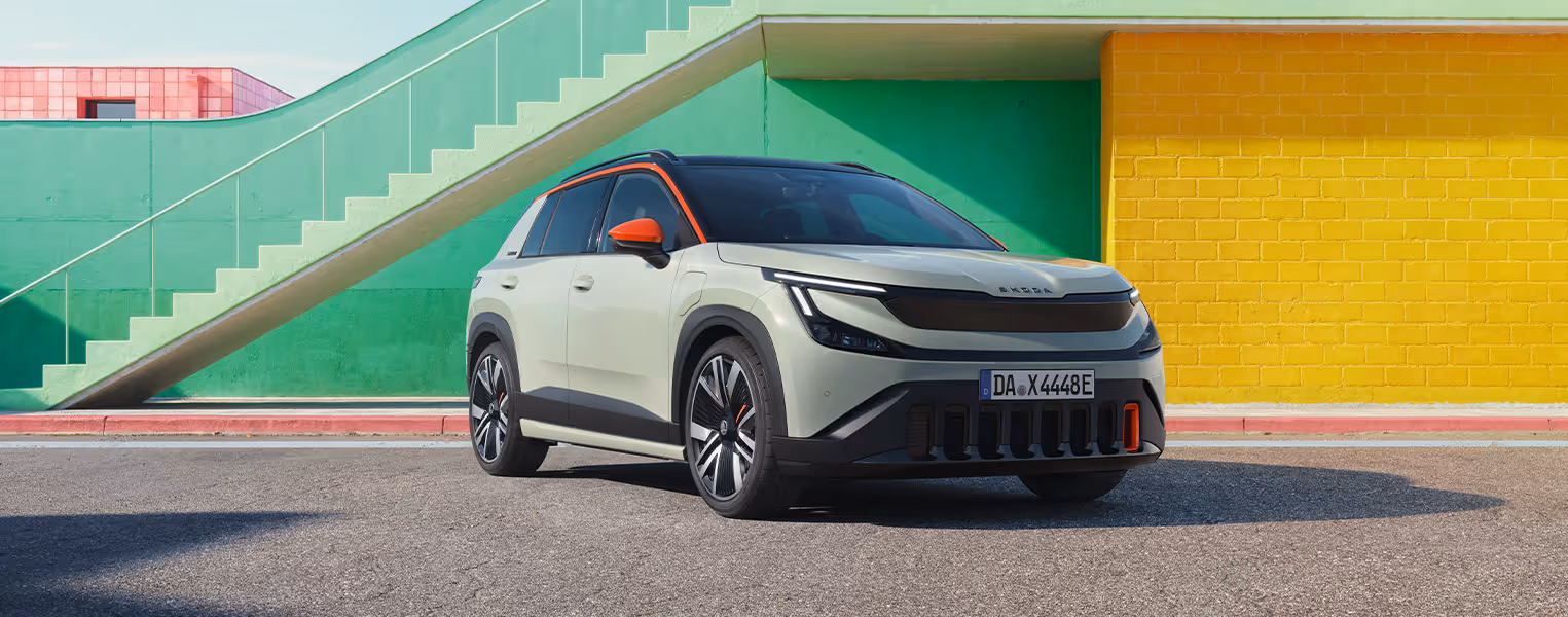 Front- und Seitenansicht des Škoda Epiq als Sondermodell "First Edition" in Platinum-Weiß Metallic, der vor einer pastellgrünen Treppe und einer gelben Wand parkt. In dieser Perspektive kommen die spezifischen orangefarbenen Akzente an der Dachlinie, den Außenspiegeln, den Leichtmetallfelgen sowie an einem vertikalen Einlass im markanten Frontstoßfänger zur Geltung. Abgerundet wird das moderne Erscheinungsbild durch die schmalen LED-Scheinwerfer und eine durchgehende schwarze Frontblende.