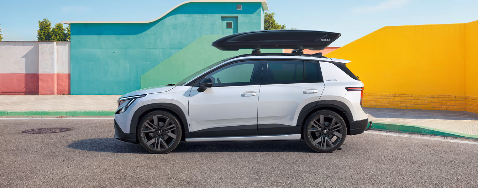 Seitenansicht des Škoda Epiq in der Farbe Platinum-Weiß Metallic, der mit einer großen, schwarzen Dachbox ausgestattet ist. Die auf den Grundträgern montierte, aerodynamische Box bietet praktischen, zusätzlichen Stauraum für Reisegepäck und Ausrüstung. Im Hintergrund hebt sich das Fahrzeug kontrastreich von geometrischen Mauern in Pastellgrün und leuchtendem Gelb ab.