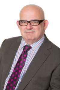 Professional headshot of bald man wearing glasses, gray blazer, and purple polka dot tie.