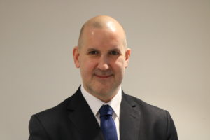 Bald man in dark suit and blue tie wearing professional headshot portrait against neutral background