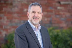 Professional headshot of middle-aged man in blazer against brick wall background