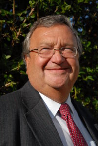 Older man wearing glasses, dark suit, and red tie smiling outdoors