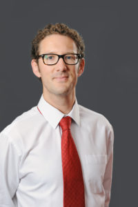 Professional headshot of man wearing white shirt and red tie against gray background