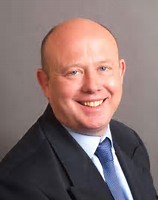 Professional headshot of a smiling bald man wearing a dark suit and blue tie.