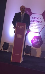 Man in suit speaking at podium with purple lighting and county signage