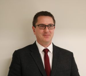 Professional headshot of a man wearing glasses, dark suit jacket, and red tie.
