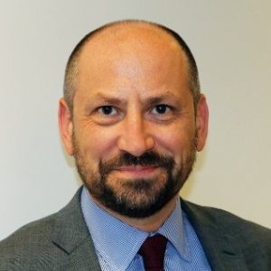 Bald man with beard wearing dark suit jacket and blue shirt.