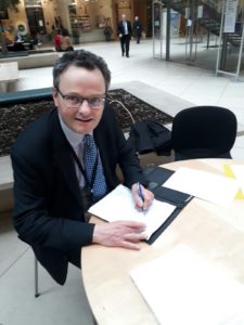 Man wearing glasses and blazer writing at table in modern building lobby.