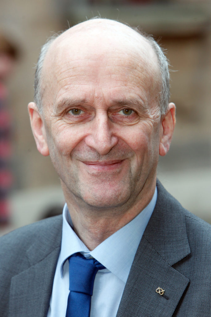 Portrait of an older man wearing a gray suit, light blue shirt, and dark blue tie.