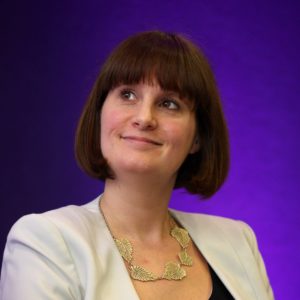 Woman with brown hair and bangs wearing white blazer against purple background