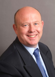 Bald man in dark suit and blue tie smiling at camera against gray background