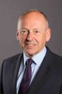 Professional headshot of a middle-aged man in business suit and tie against gray background.