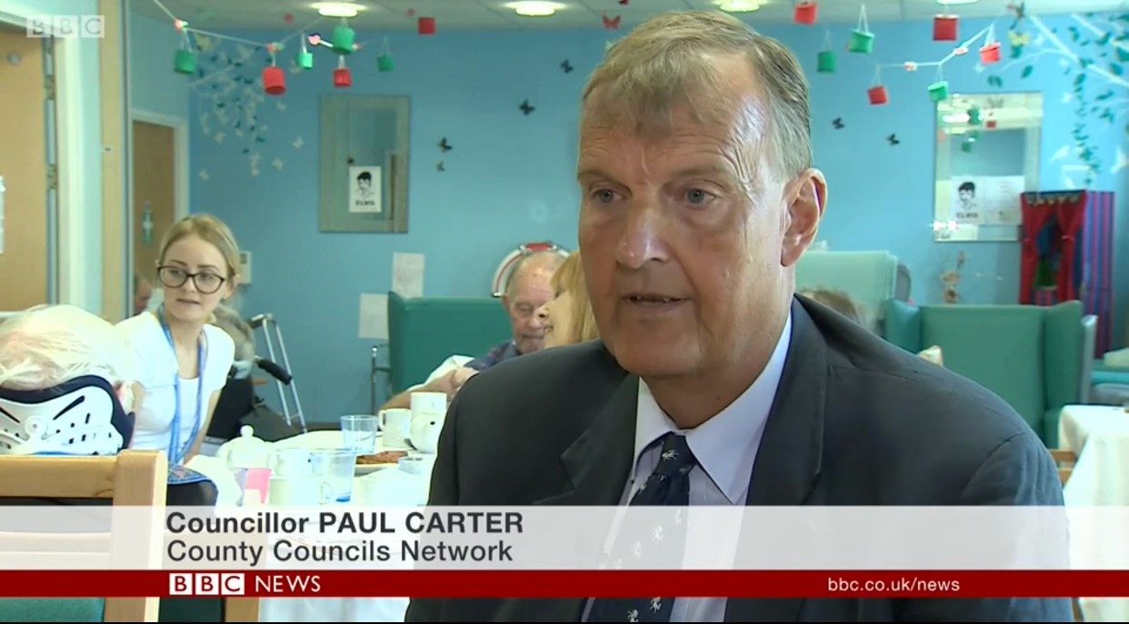 Councillor Paul Carter speaks at desk on BBC News with colleagues in decorated meeting room background