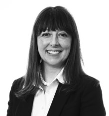 Professional headshot of woman with long dark hair and bangs, wearing dark blazer.