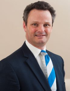 Professional headshot of a man in dark suit and blue tie against beige background