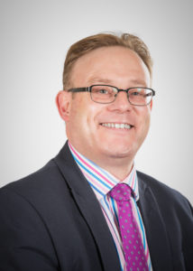 Professional headshot of middle-aged man wearing glasses, dark blazer, and purple tie.