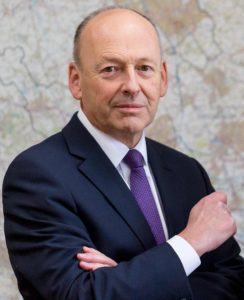 Bald man in dark suit and purple tie, arms crossed, professional portrait