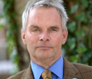 Middle-aged man with gray hair wearing blue shirt and brown jacket outdoors.