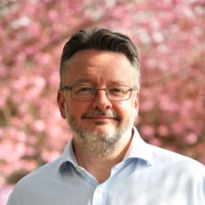 Man wearing glasses and white shirt smiling in front of pink flowers